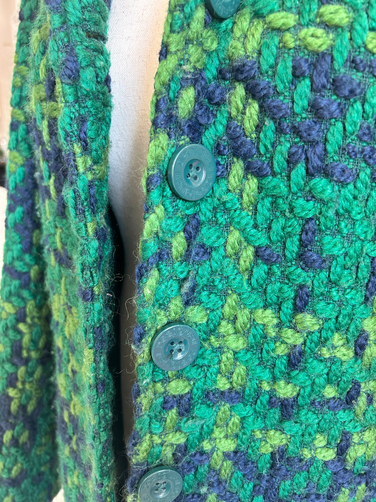 Close-up of a textured knit cardigan featuring a green and navy blue leaf-like pattern, with dark green buttons, displayed on a cream-colored dress form.