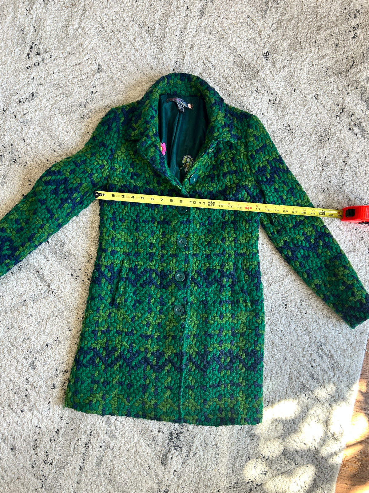 A green and blue patterned wool coat is laid flat on a textured rug with a yellow tape measure stretched across the chest to show the width. 