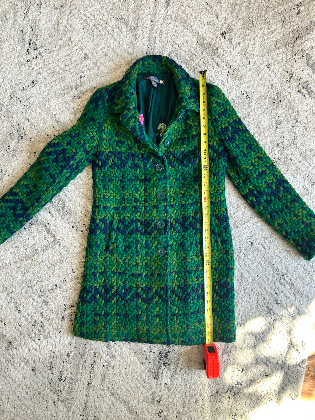 A green and blue patterned wool coat is laid flat on a textured white rug, with a yellow tape measure standing vertically next to it for size reference.