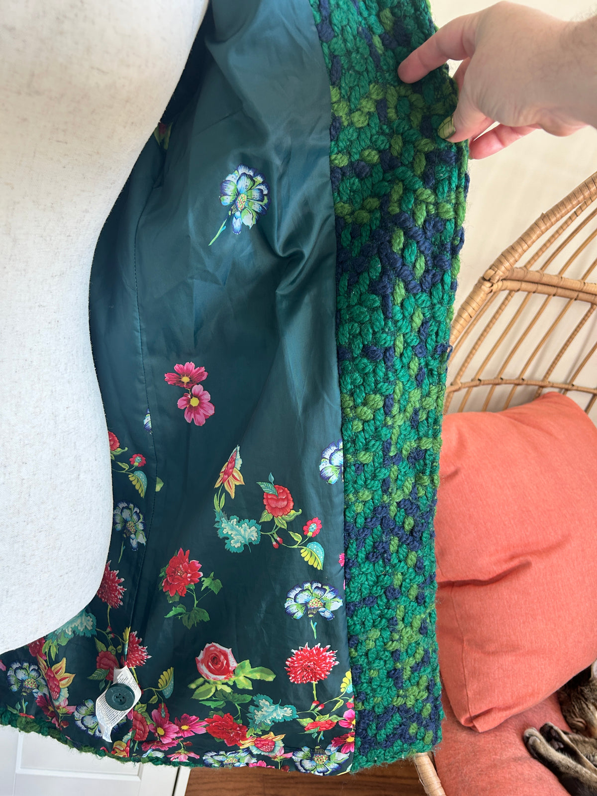 A close-up of a garment or fabric panel draped on a mannequin, showing a dark teal satin-like interior with a colorful floral print and a textured green-and-blue patterned border. A hand is holding the edge, and part of a wicker chair with a coral pillow is visible in the background.