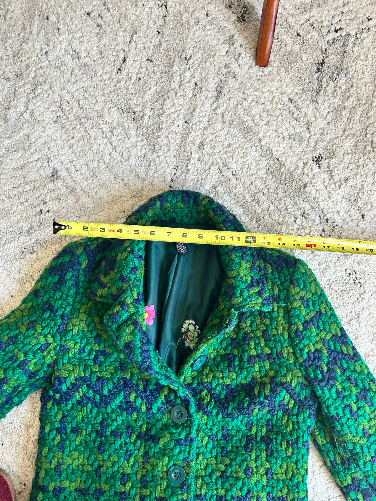 A green and purple marled tweed blazer is laid flat on a beige carpet, with a yellow tape measure stretched horizontally across the chest to show the size.
