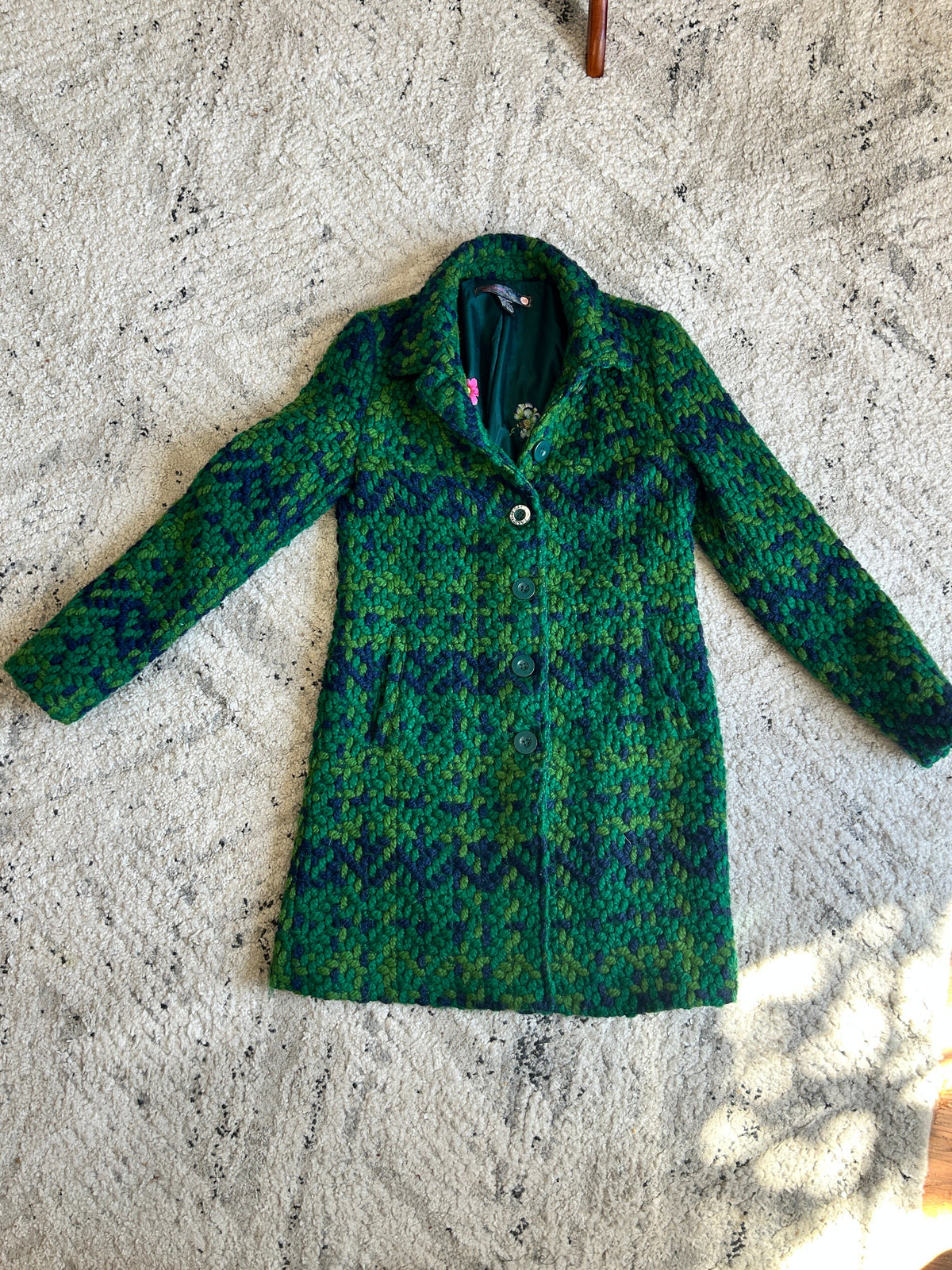 A green and blue patterned wool coat  laid flat on a textured cream rug.