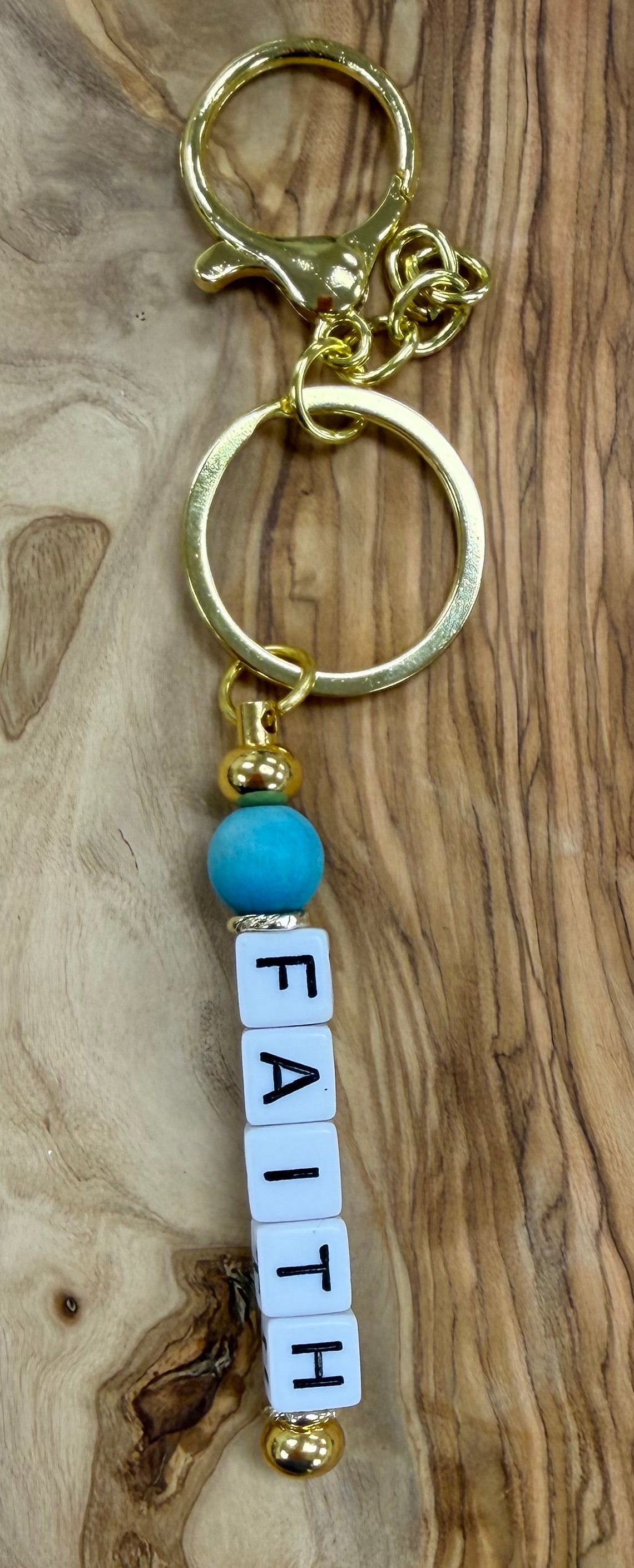 Gold Keychain with 'FAITH' letter beads  with a blue accent bead, on a wooden surface