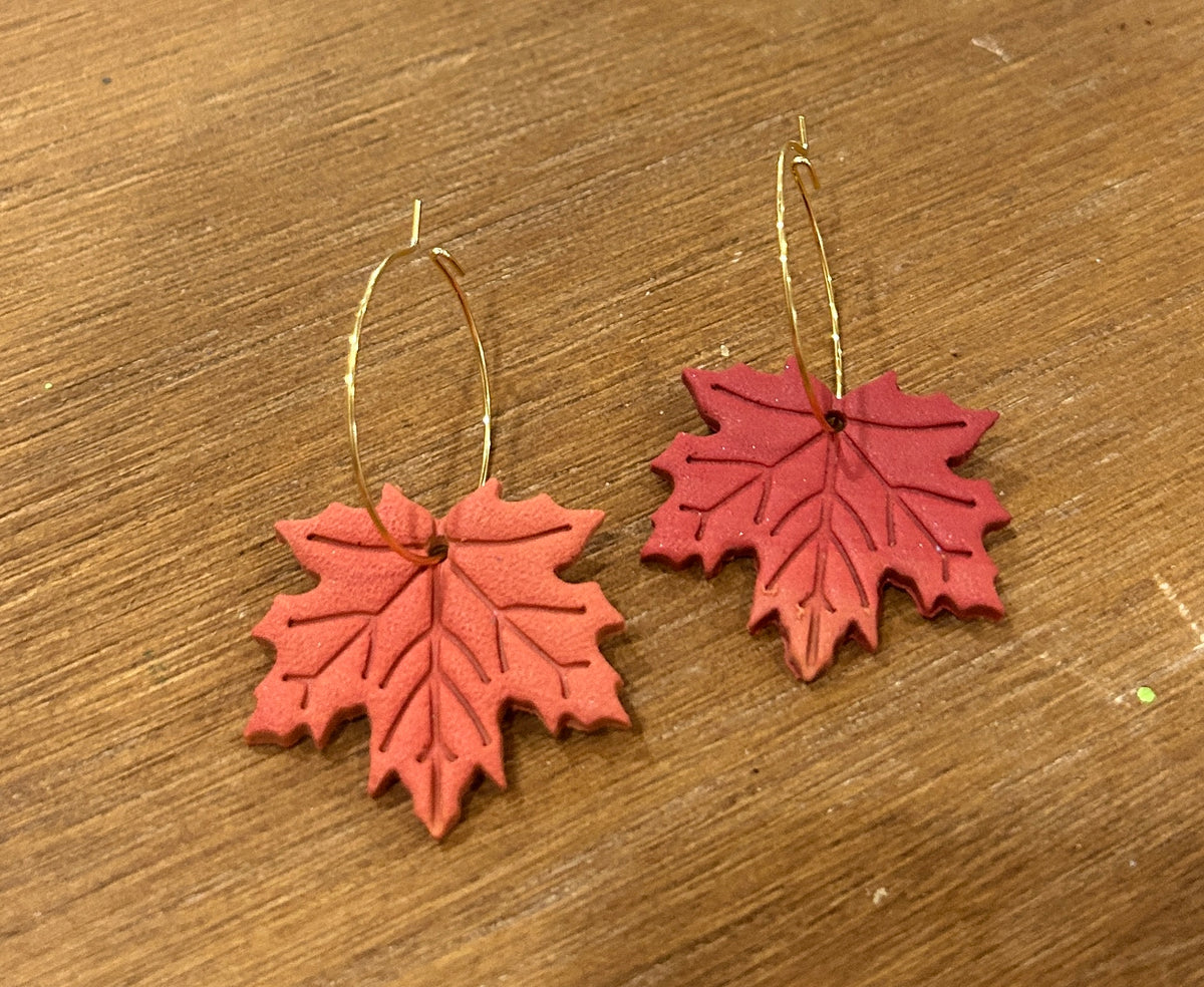 A pair of handmade maple leaf earrings with a red-to-orange gradient and gold hoops displayed on a wooden surface.