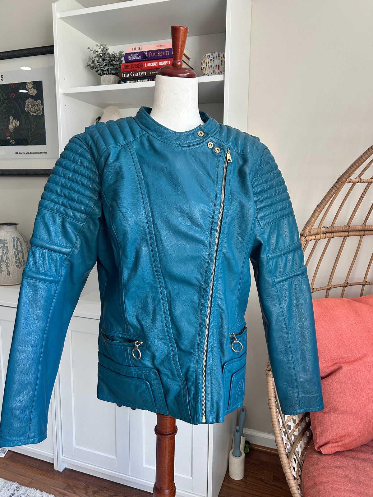 A teal blue leather jacket with a diagonal zipper and quilted shoulder panels, displayed on a white mannequin torso in a home setting with a bookshelf and rattan chair visible in the background.