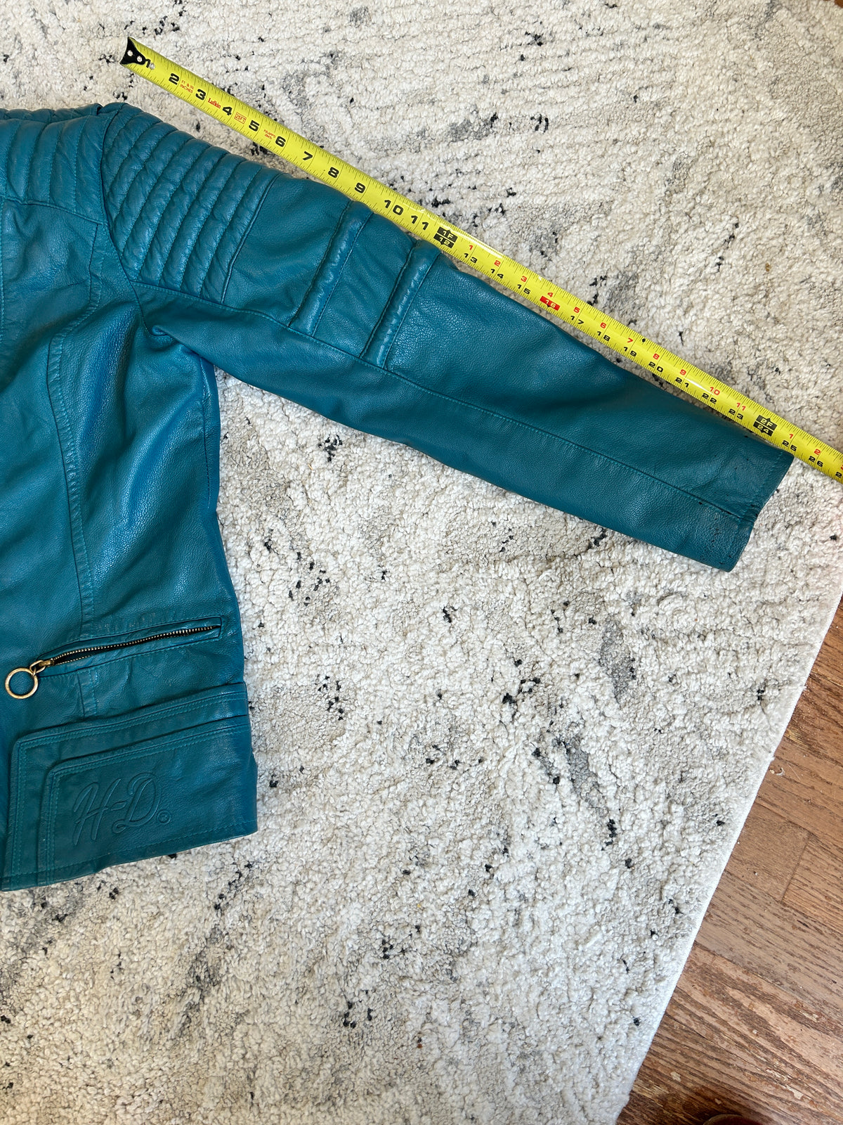 A teal blue leather jacket laid flat on a textured light gray rug, with a yellow tape measure placed diagonally across the upper portion to indicate size.