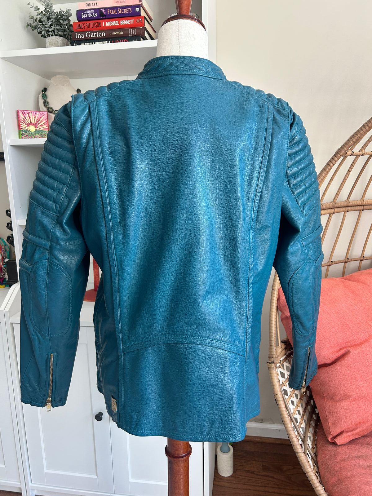 A teal blue leather motorcycle jacket displayed on a mannequin torso, showing the back view with quilted sleeve details and zippered pockets, set against a home interior background.