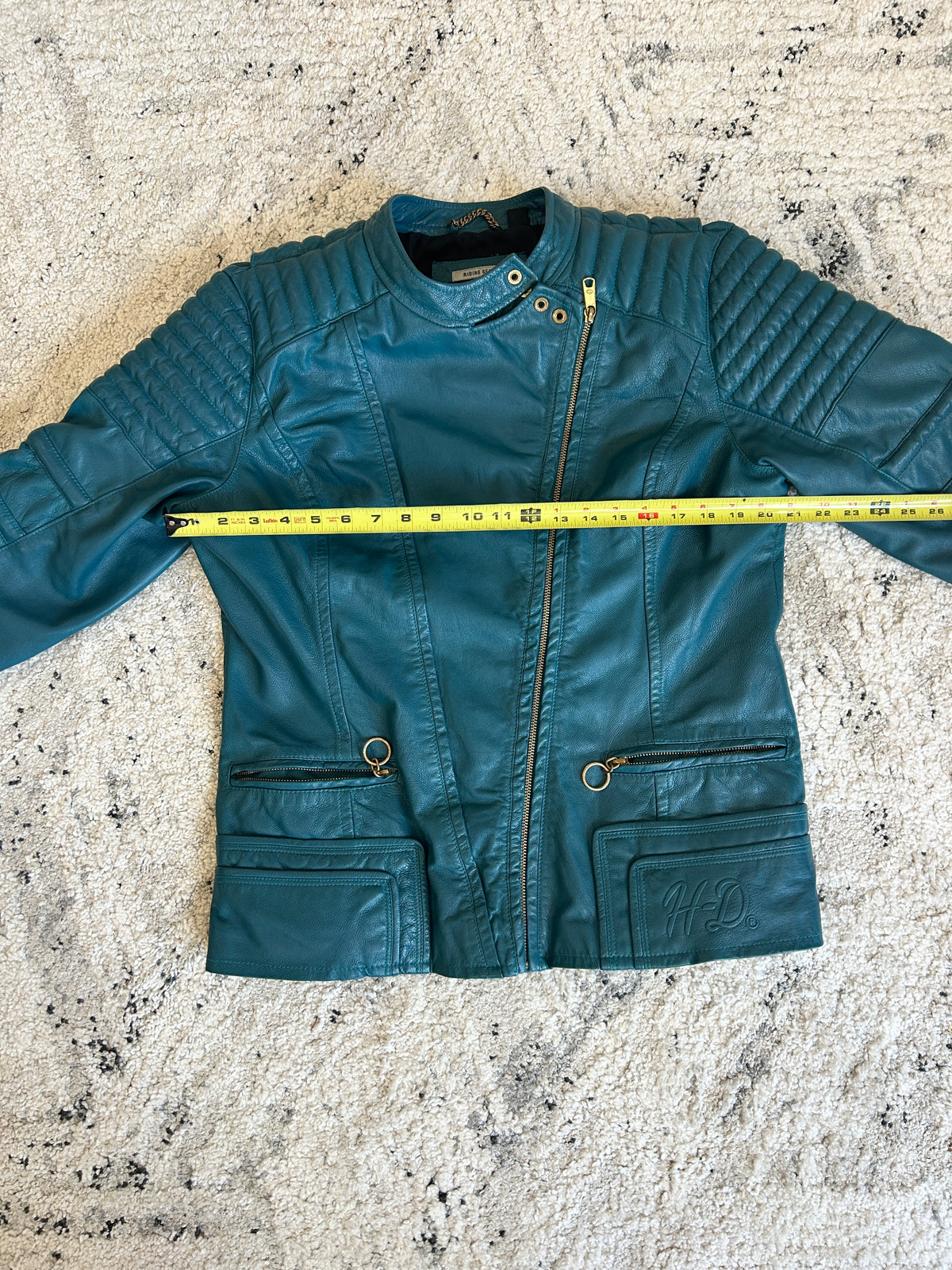 Teal leather jacket on a textured white surface with yellow measuring tape showing chest width.