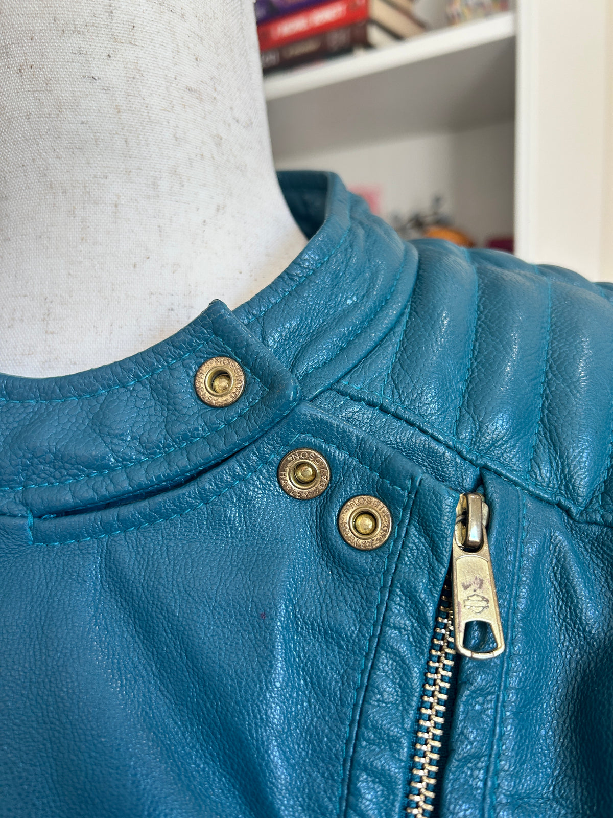 Close up view of a teal leather jacket with asymmetrical gold-toned zipper and snaps