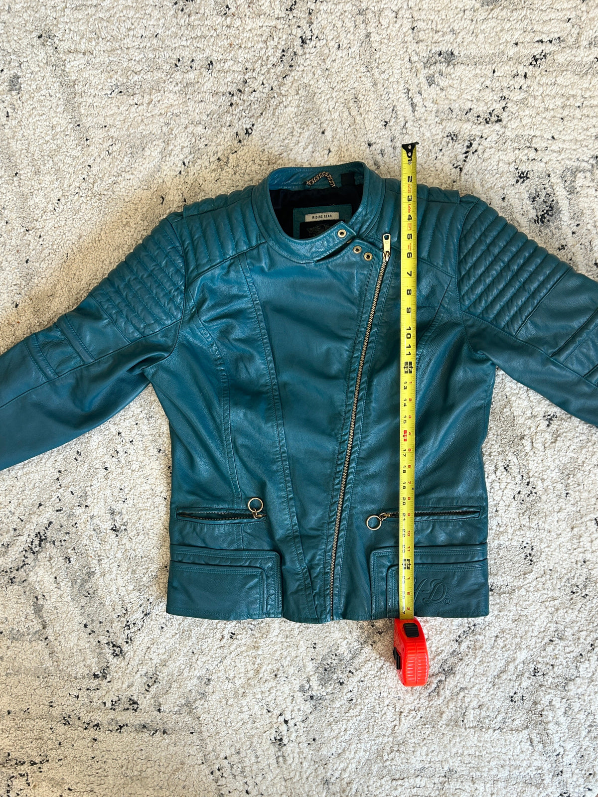 Teal leather jacket on textured white rug with yellow measuring tape showing length