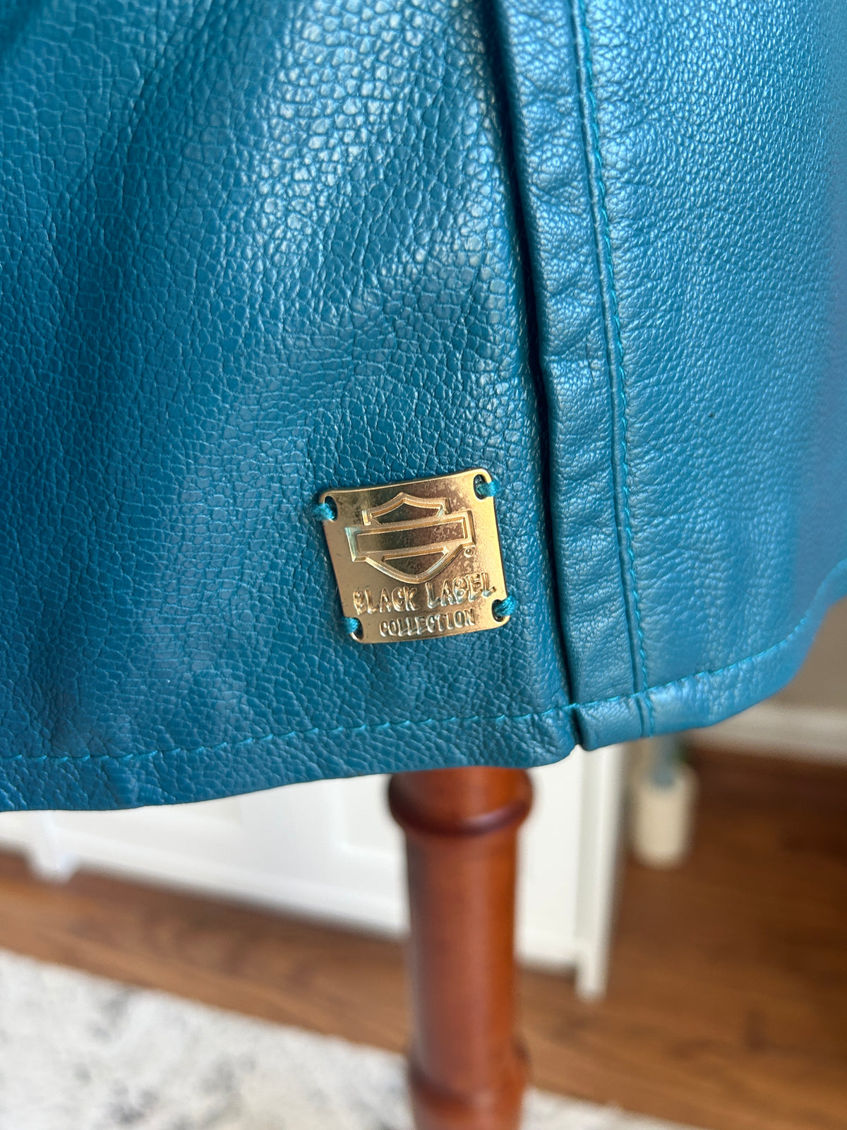Close-up of a teal blue pebbled leather jacket with a gold-tone Harley-Davidson Black Label Collection metal badge, resting on a wooden stand.