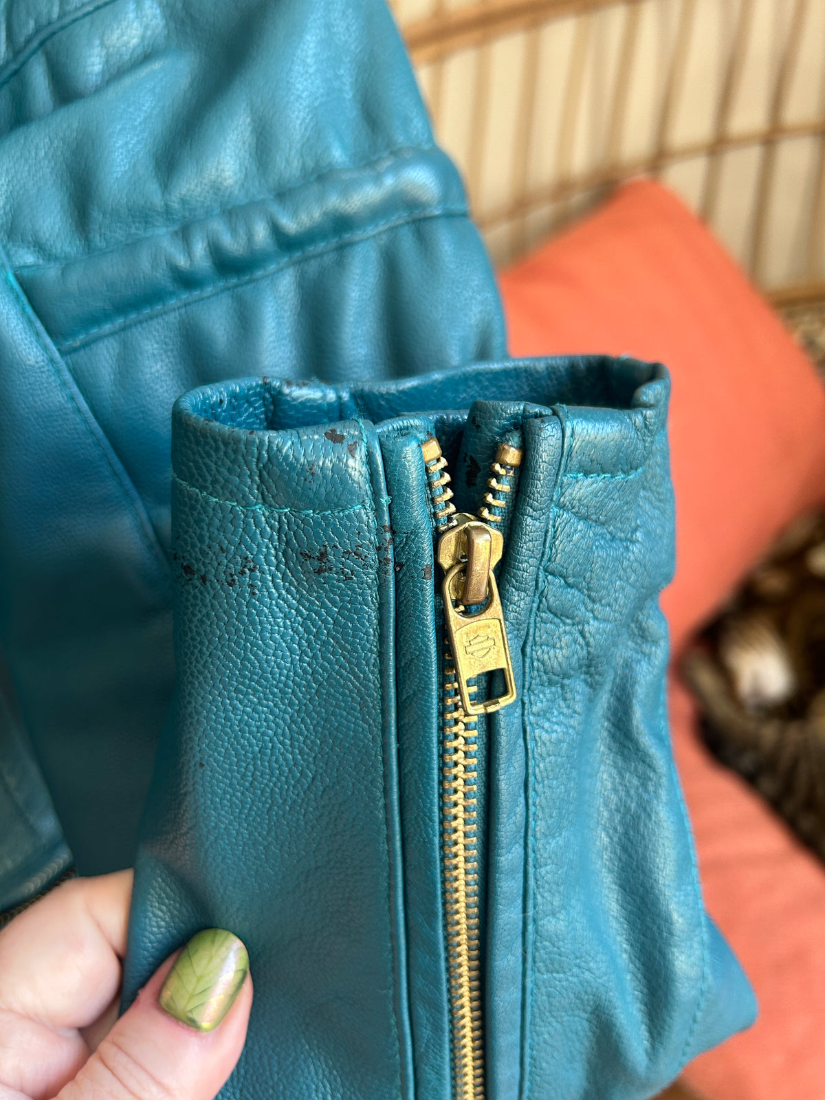 Close up view of gold-toned zipper on the wrist of a teal leather jacket. Slight color loss markings are visible.