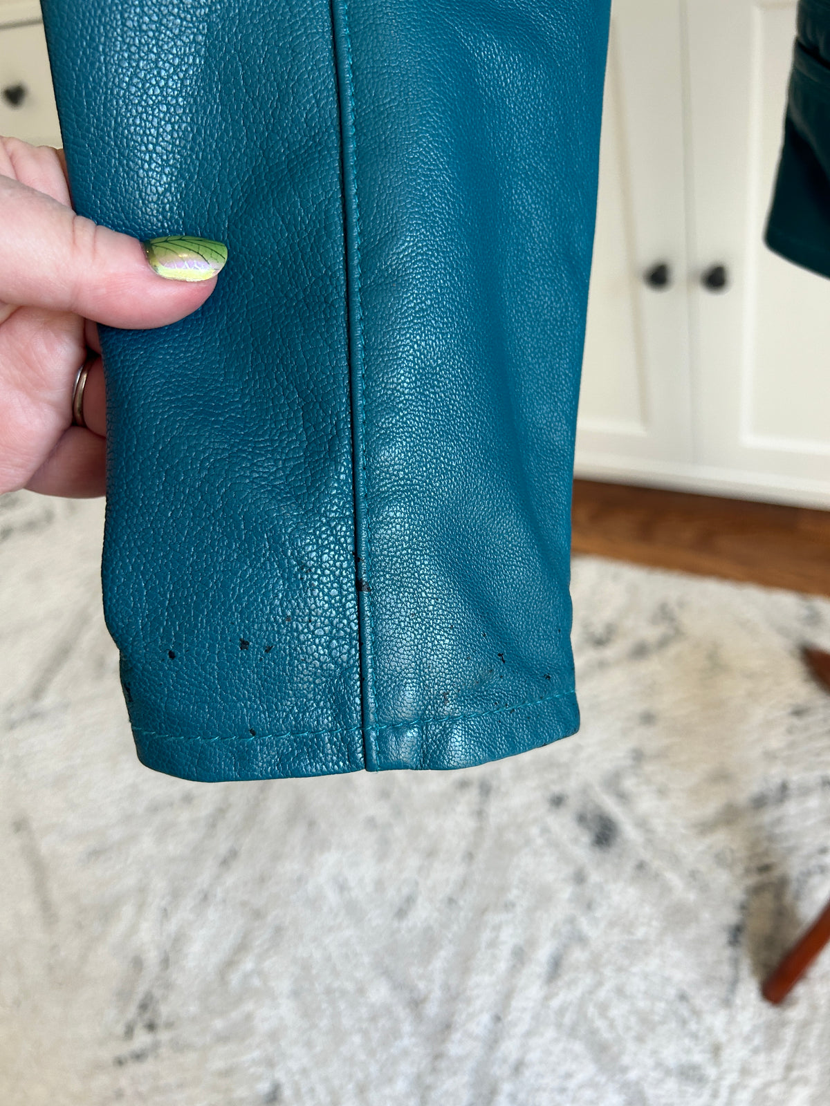 A close-up of a teal blue leather sleeve being held by a hand, showing the texture and slight color loss with a home interior background.