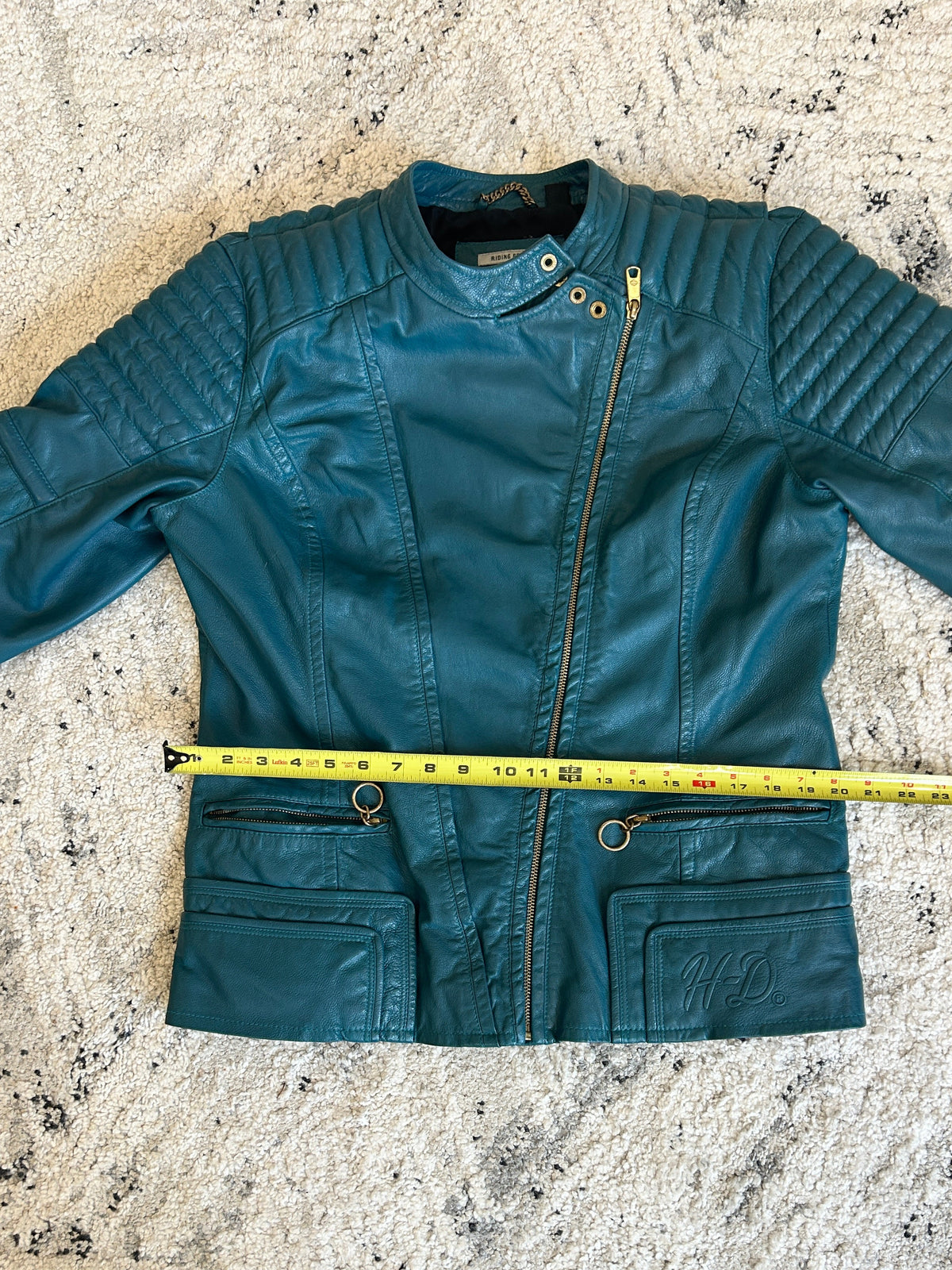 A teal blue leather jacket with a front zipper and flap pockets, laid flat on a textured beige rug with a yellow tape measure spanning across the chest to show width.