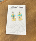A pair of handmade dangle earrings by Juniper Designs, featuring an alien abduction scene with a cow