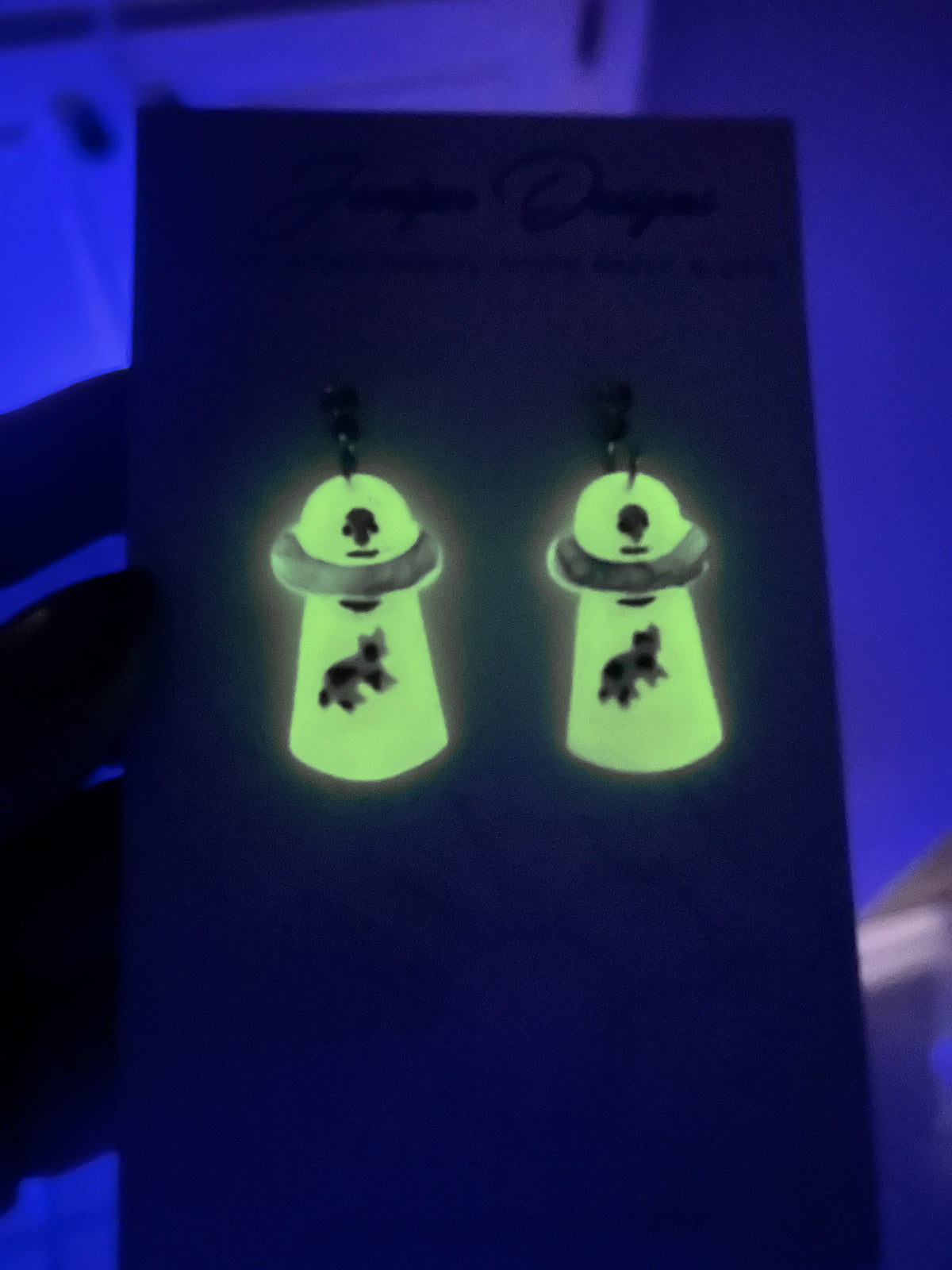 Glow-in-the-dark dangling alien abduction earrings photographed in the dark under blue light. The earrings appear bright neon green and feature small black graphic details.