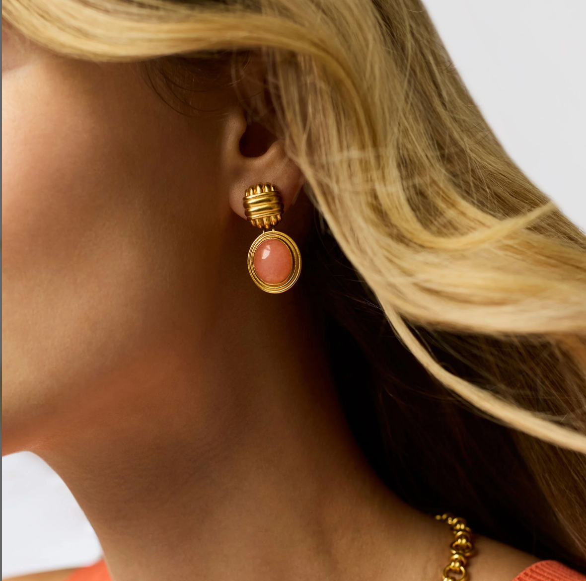 A woman with blonde hair wearing a gold stud earring with a coral colored jade dangle.