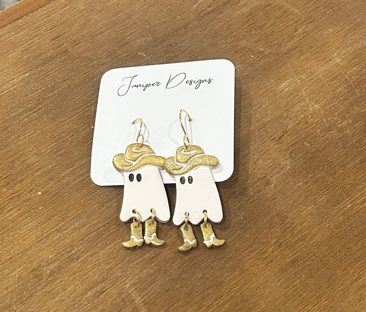 A pair of white ghost-shaped dangle earrings with gold cowboy hats and boots, displayed on a white card labeled 'Junoiper Designs' on a wooden surface.