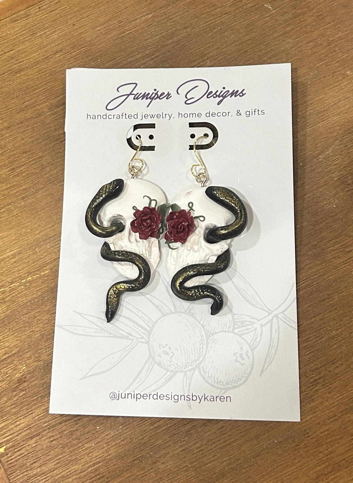 A pair of skull earrings featuring snakes and roses earrings displayed on a white card from Juniper Designs, placed on a wooden surface. 