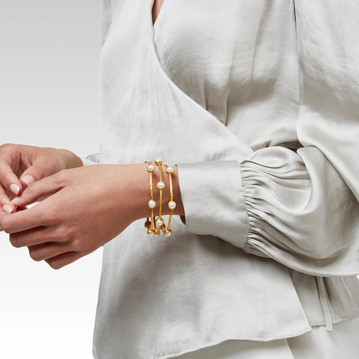 Person wearing three gold bracelets with pearls on a plain background