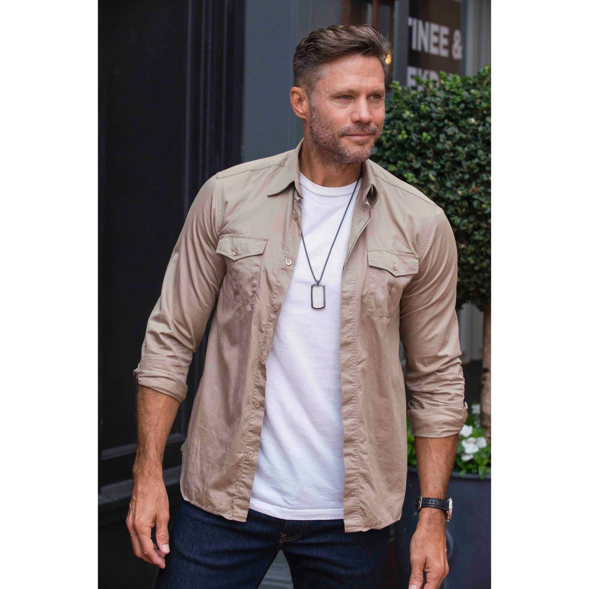 Man in a beige shirt over a white t-shirt models a gunmetal necklace with a dog tag-shaped monocle pendant.