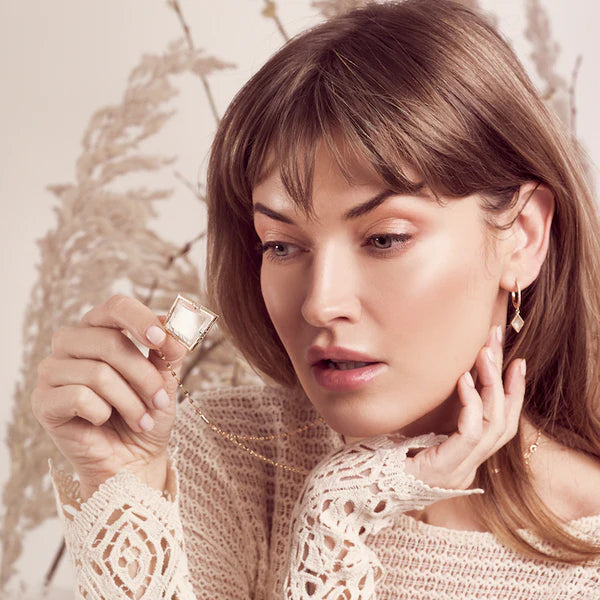 Woman in a white top looking thru a diamond-shaped monocle pendant on a silver necklace.
