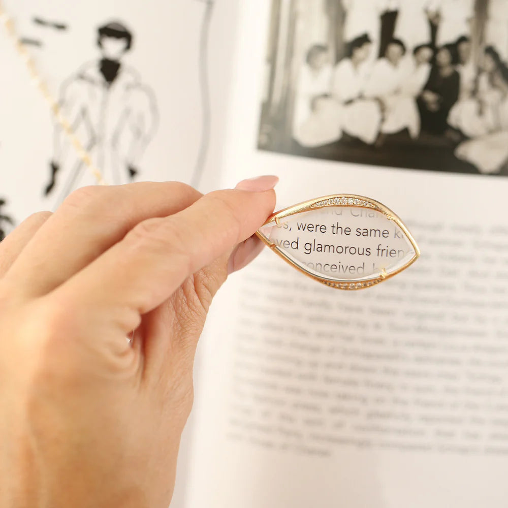 Woman using a diamond accented teardrop-shaped monocle pendant to clearly read small text in a book.