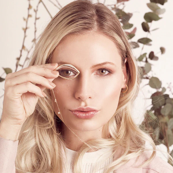 Woman looking thru a teardrop-shaped monocle pendant on a gold necklace with a natural background