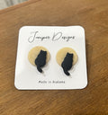 A pair of stud earrings by Juniper Skies, featuring black cat silhouettes sitting on light beige circular backgrounds, mounted on a white display card that reads 'Juniper Designs' and 'Made in Alabama', placed on a wooden surface.