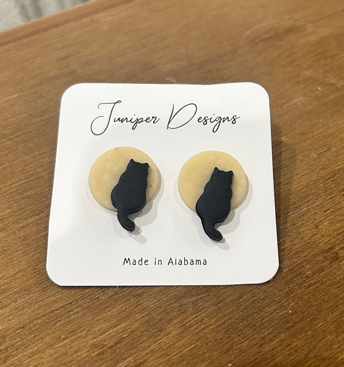 A pair of stud earrings by Juniper Skies, featuring black cat silhouettes sitting on light beige circular backgrounds, mounted on a white display card that reads 'Juniper Designs' and 'Made in Alabama', placed on a wooden surface.