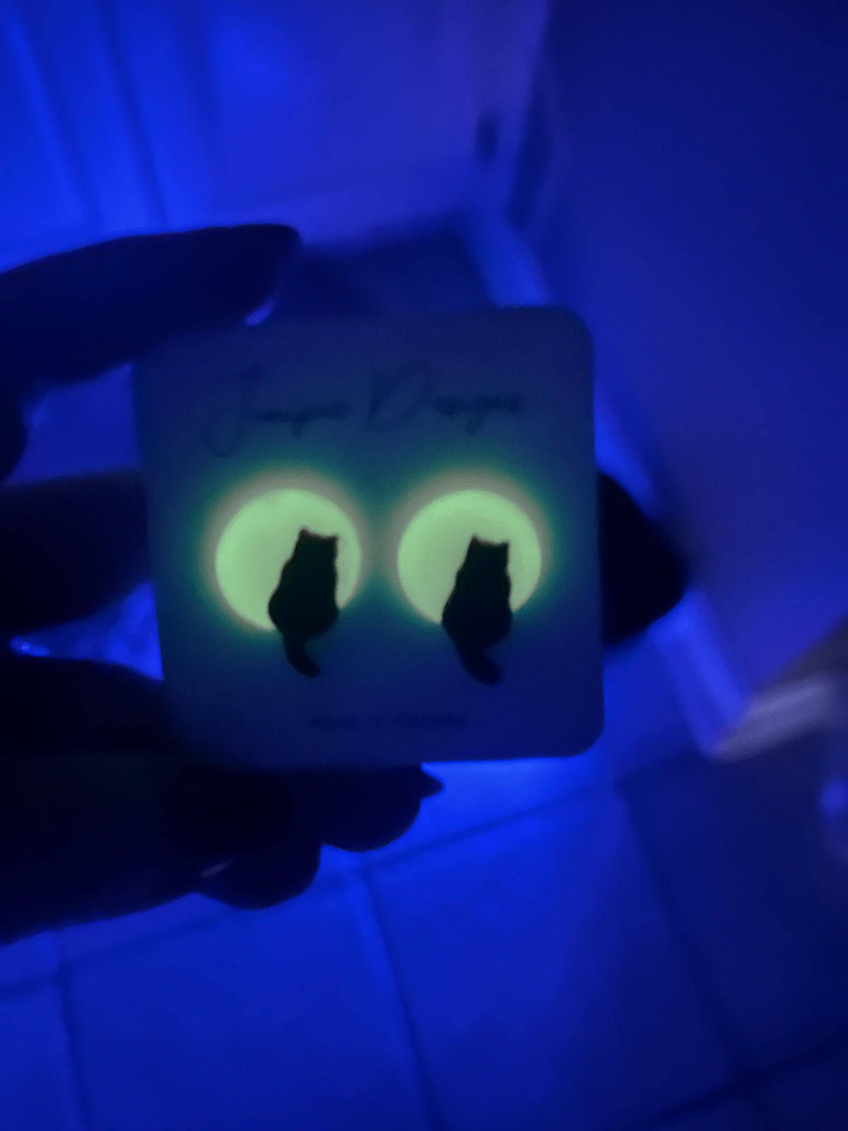 A hand holds a square card featuring a pair of glowing green cat-shaped earrings on a dark, blue-lit background. The card has 'Jupiler Designs' printed on it.