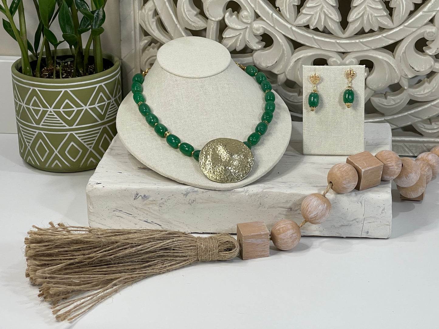 The necklace is made of rich, glossy green oval beads, each separated by small gold-toned spacer beads. At the center of the necklace is a large, round, hammered gold pendant—bold and textured with an organic, artisan-crafted look. The necklace is displayed on a slightly reclined cream-colored stand, giving it a casual, elegant presentation.

The matching earrings hang on a small cream-colored earring display. Each earring has a single green oval bead—matching the necklace—suspended from a delicate gold-ton