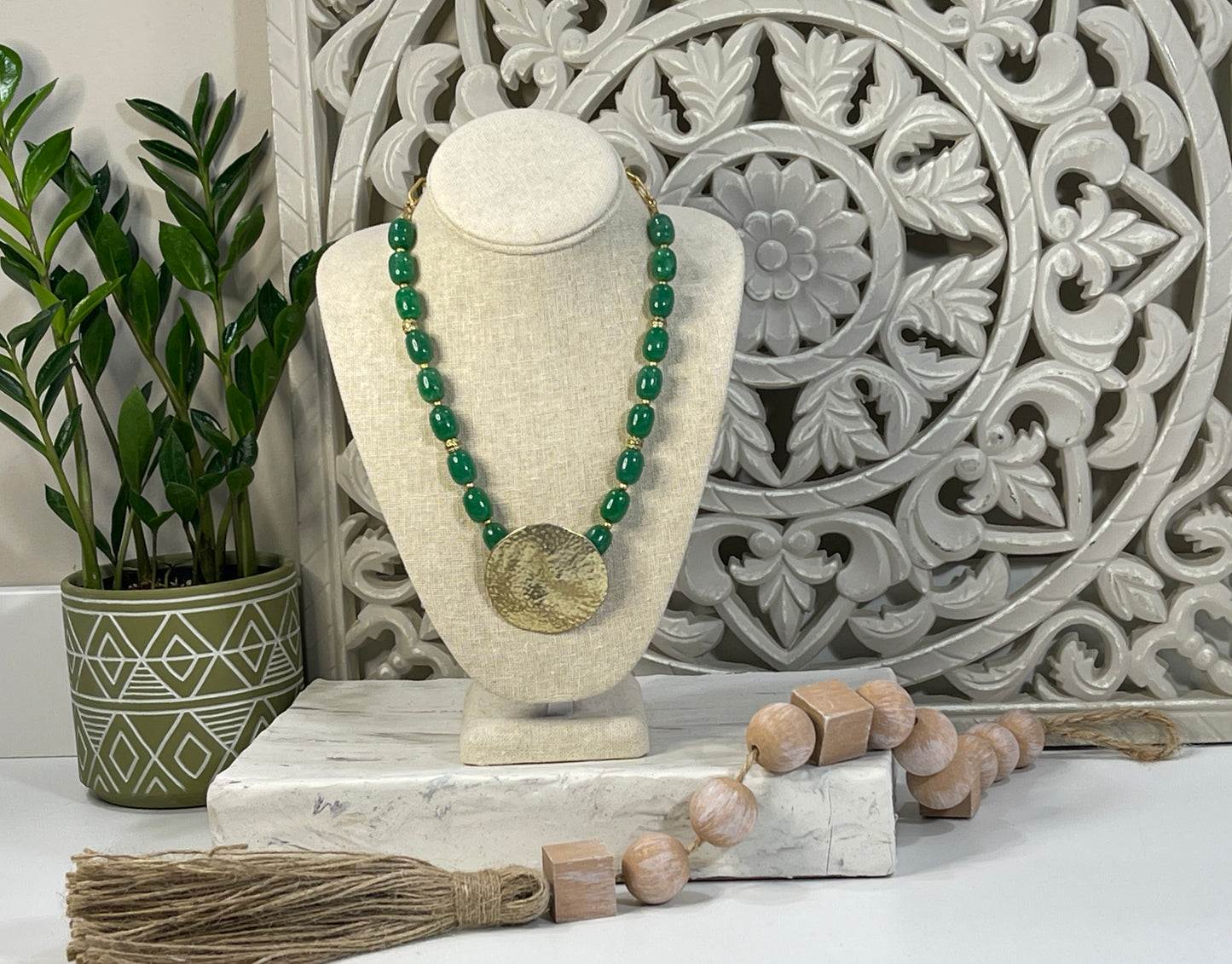 This photo shows a beautifully styled jewelry display. The necklace features bold, glossy green oval beads strung symmetrically, leading to a large, round, hammered gold pendant that hangs prominently at the center.