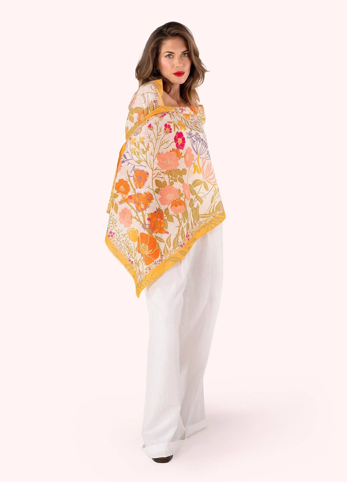 Woman wearing a orange, yellow and white floral scarf and white pants on a white background
