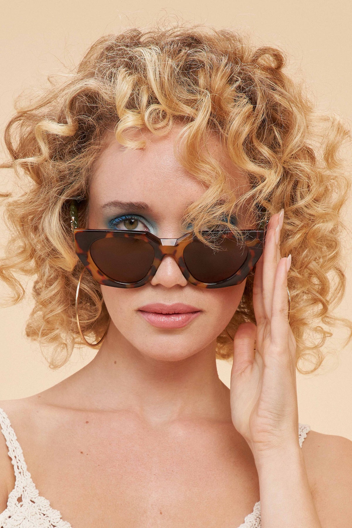 Woman with curly blonde hair wearing olive tortoiseshell sunglasses against a beige background