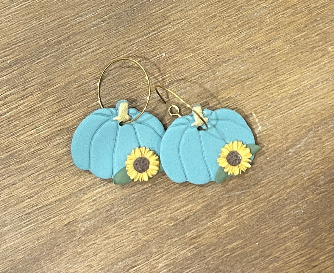 A pair of handmade hoop earrings featuring a sage pumpkin decorated with yellow sunflowers, displayed on a wooden surface.