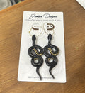 A pair of black snake drop earrings with gold accents, displayed on a white card from Juniper Designs, placed on a wooden table.