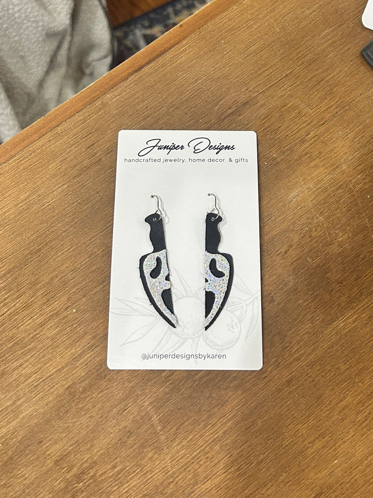 A pair of knife-shaped drop earrings on a white display card, placed on a wooden table. The knives have a stabby scream face on them.