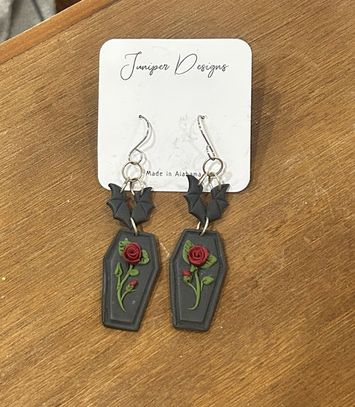 A pair of gothic coffin-shaped dangle earrings with red roses and green leaves, displayed on a white card labeled 'Juniper Designs' on a wooden table.