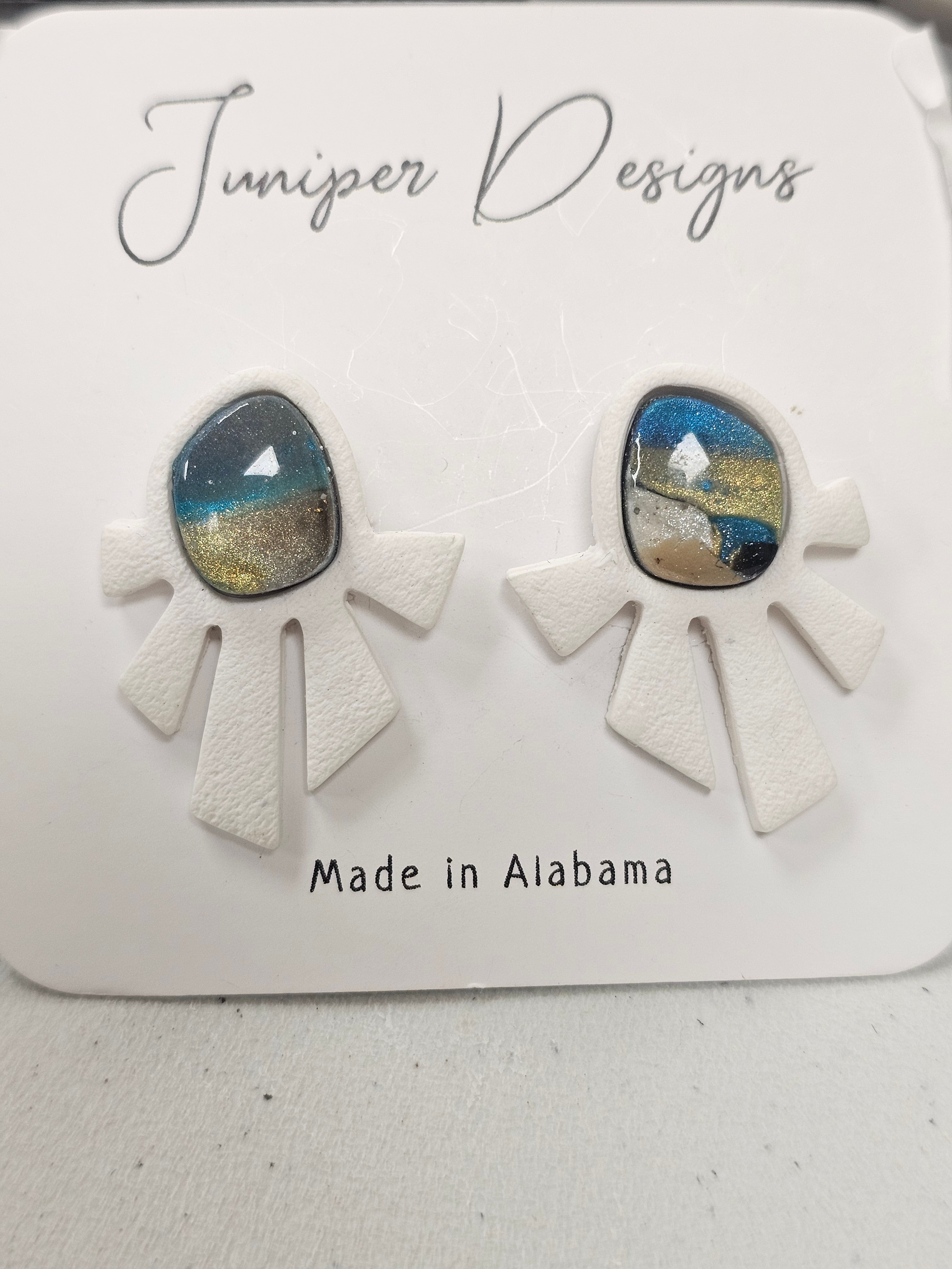 Pair of earrings with white geometric shapes and blue and gold center on a branded card.