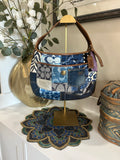 Blue, brown, and purple patchwork bag hanging on a gold purse display on a white shelf with home decor