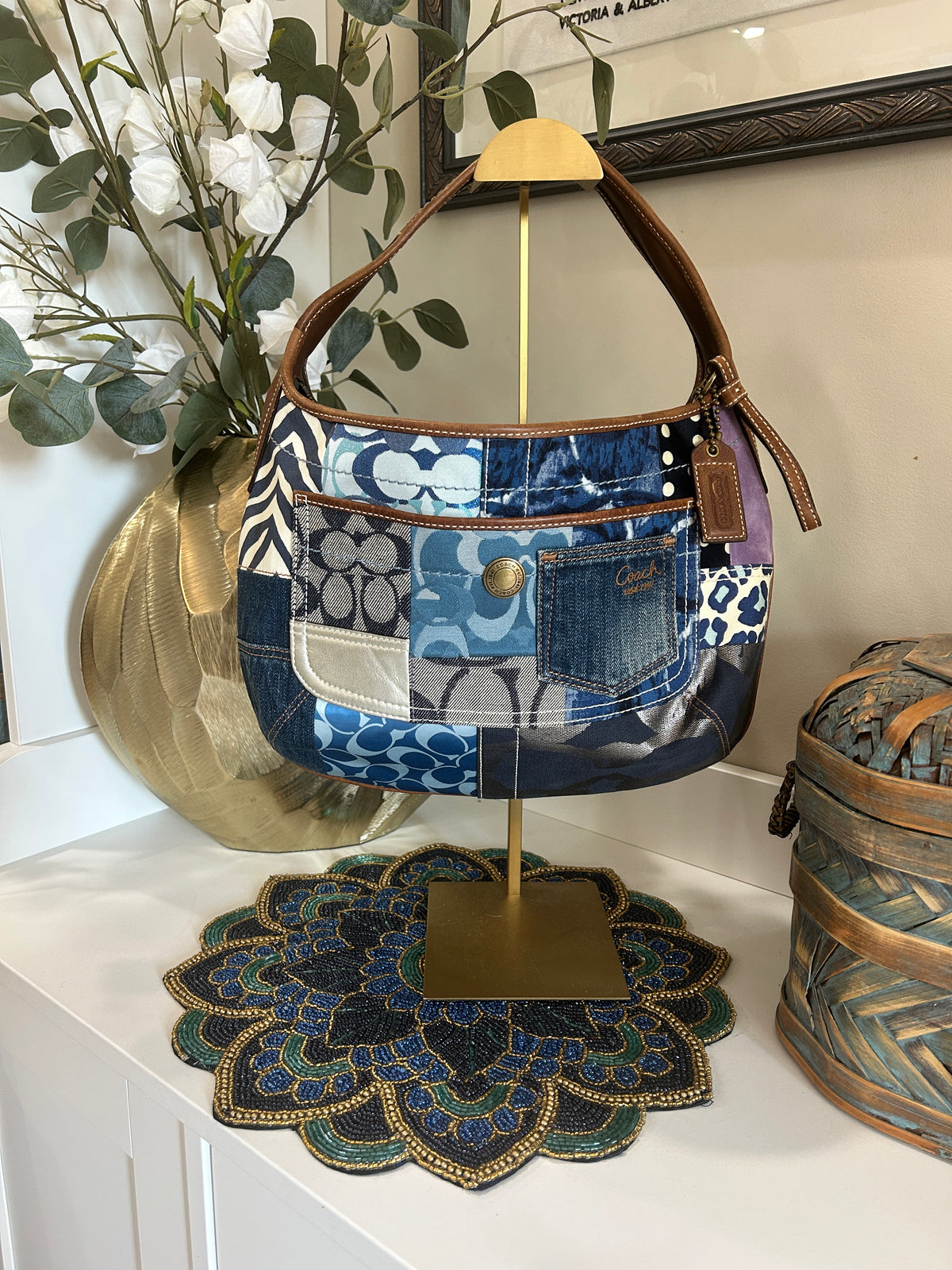 Blue, brown, and purple patchwork bag hanging on a gold purse display on a white shelf with home decor