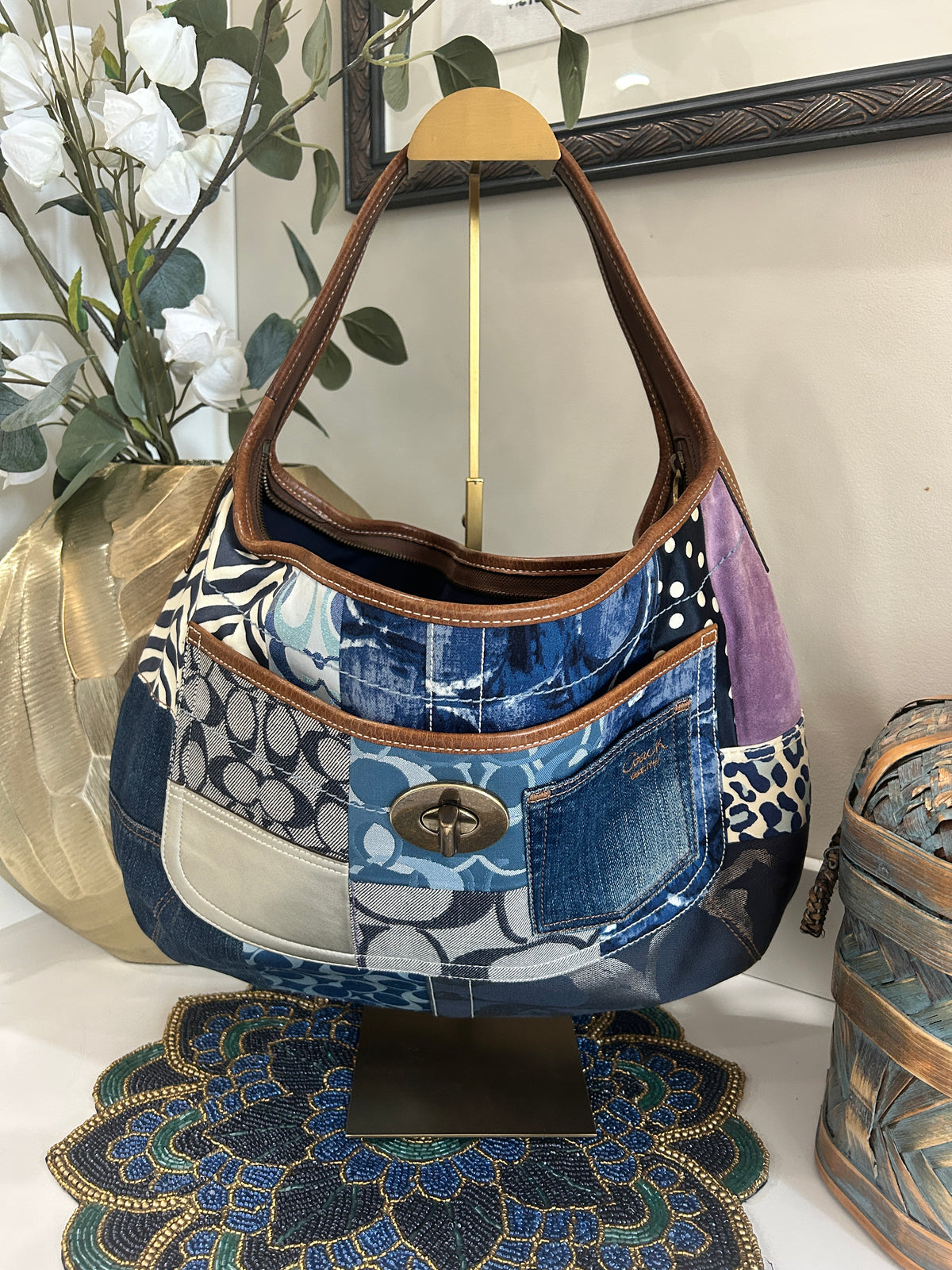 Blue, brown, and purple patchwork bag hanging on a gold purse display on a white shelf with home decor