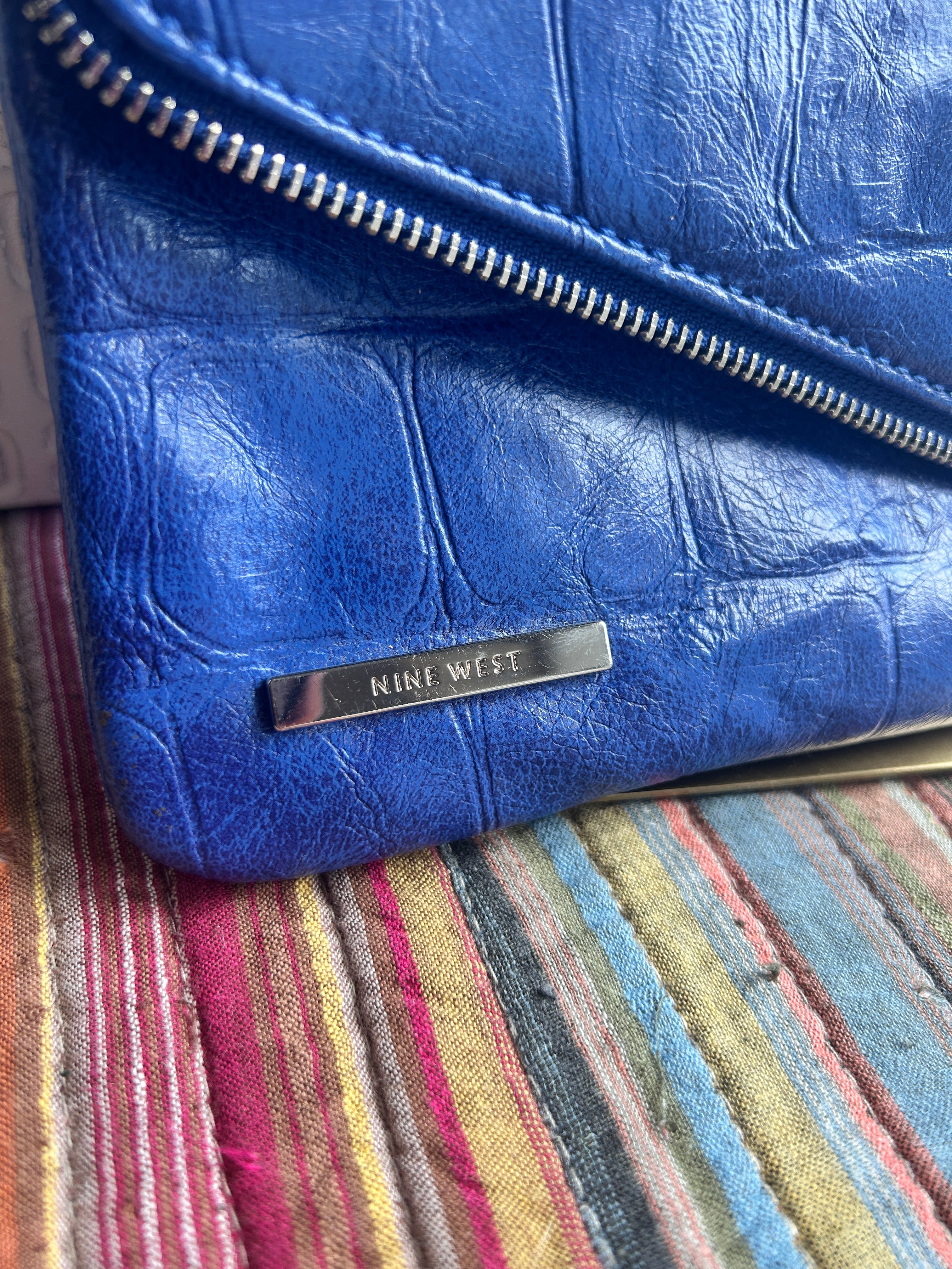 A close-up of a blue textured leather clutch or wallet with a silver zipper and a vertical silver metal brand plaque engraved with 'Nine West." The product is shown resting on a striped fabric surface.
