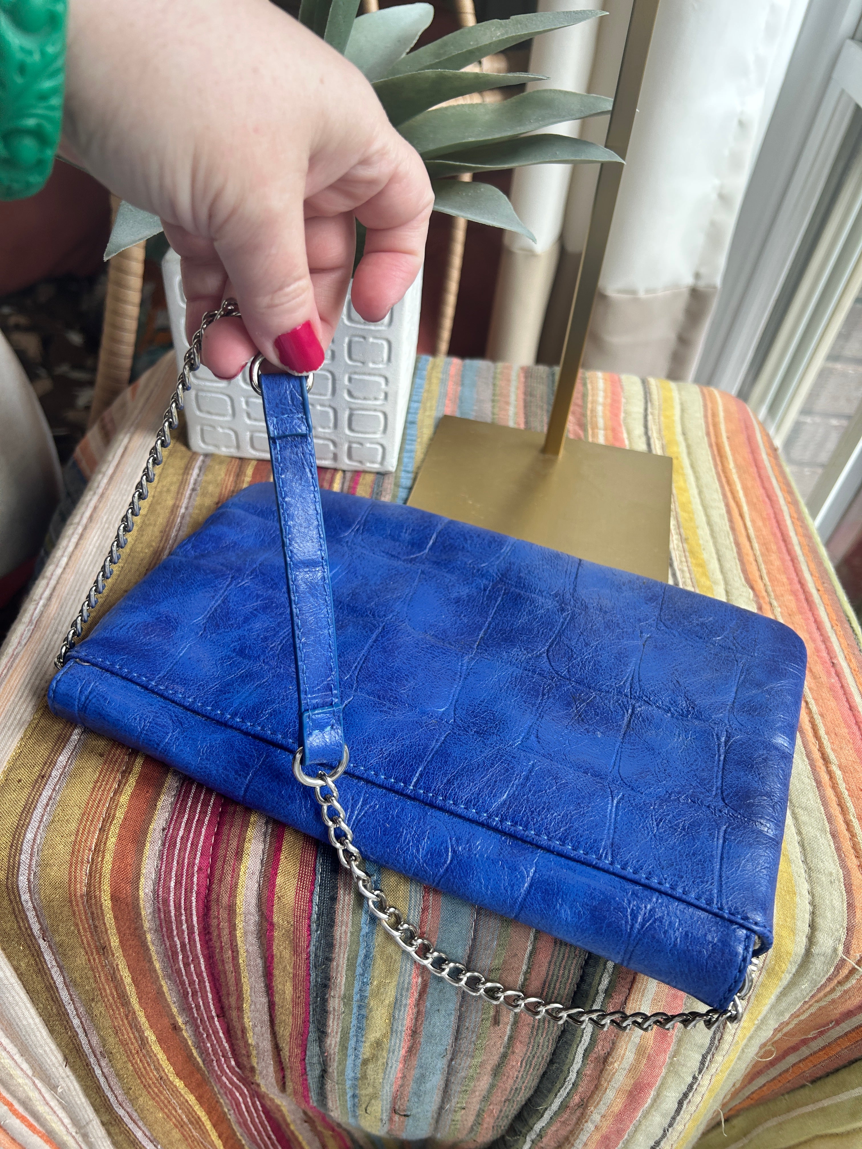A blue crocodile-embossed leather clutch bag with a silver chain strap, held by a hand with red nail polish against a striped fabric background.