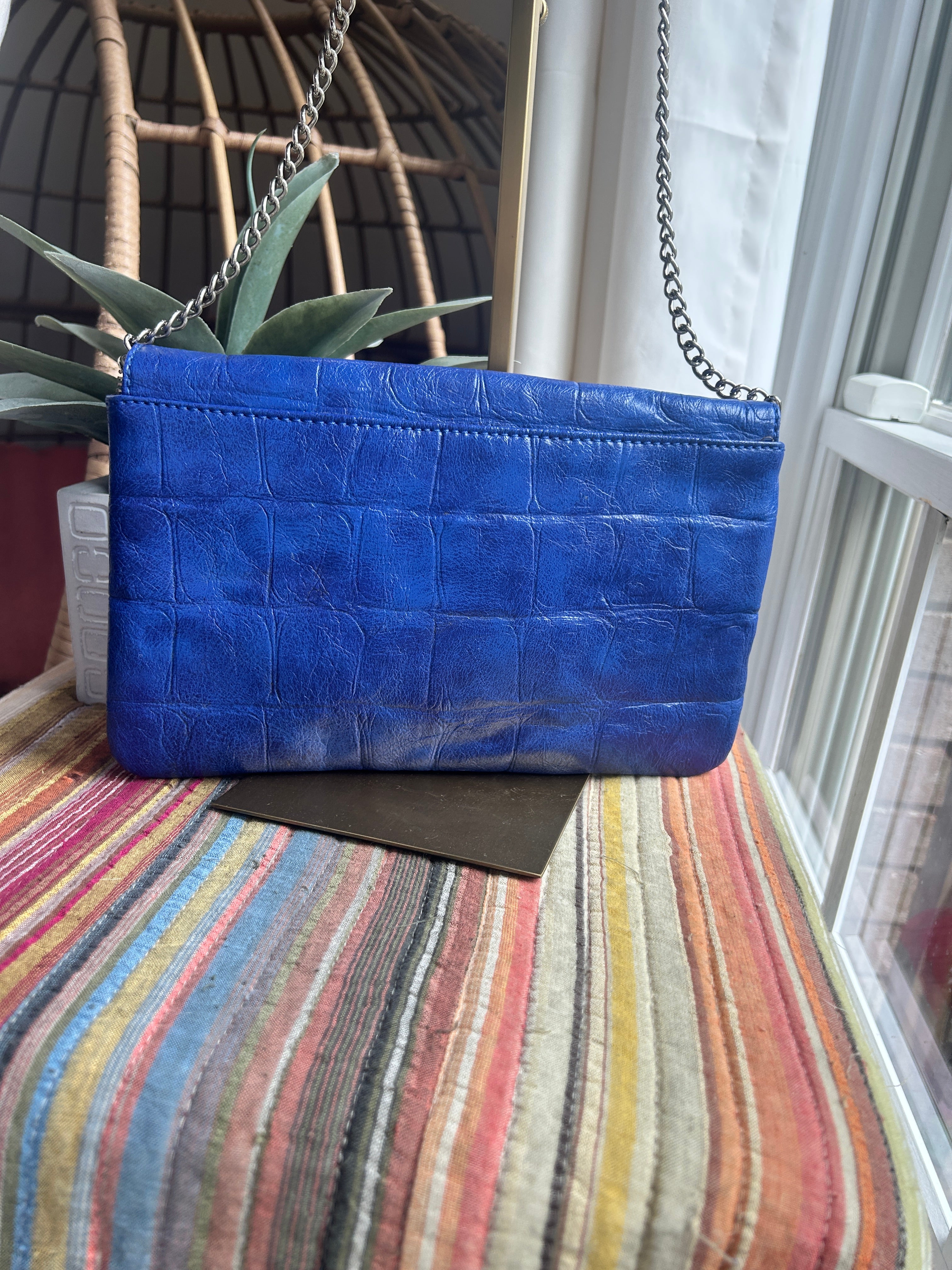 A blue crocodile-embossed leather clutch bag with a silver chain strap, resting on a striped multicolor blanket.