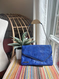 A blue crocodile-embossed leather clutch bag with a silver chain strap, resting on a striped blanket next to a potted plant.